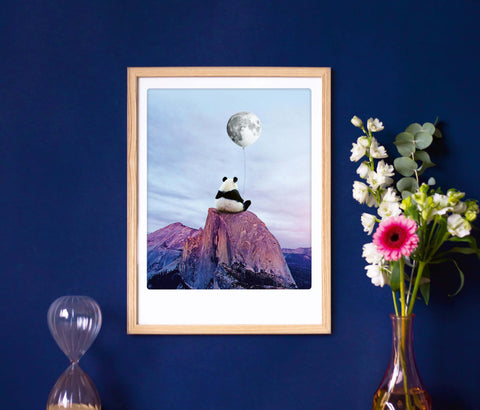 Gerahmtes Kunstwerk eines Pandas auf einem Berg, der einen Mondballon hält, aufgehängt an einer blauen Wand neben Blumen und einer Sanduhr.