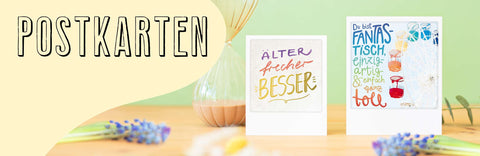 Zwei bunte Postkarten auf einem Holztisch mit Blumen und dem Wort POSTKARTEN in großer Schrift auf der linken Seite.