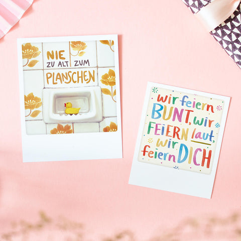 Zwei bunte Karten auf rosafarbener Oberfläche; auf der einen ist eine Gummiente abgebildet, auf der anderen ein deutscher Festtext.
