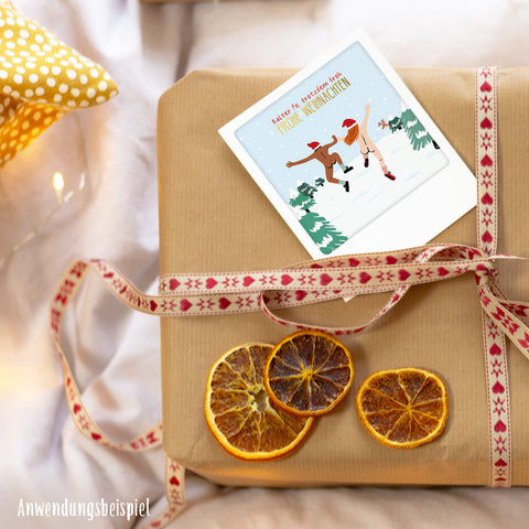 Ein eingepacktes Geschenk mit getrockneten Orangenscheiben, festlichem Band und einem Pickmotion Besinnliche Momente | 5er Kleine Postkarten-Set mit Eisläufern und weihnachtlichen Sprüchen in Goldeffekten oben drauf.