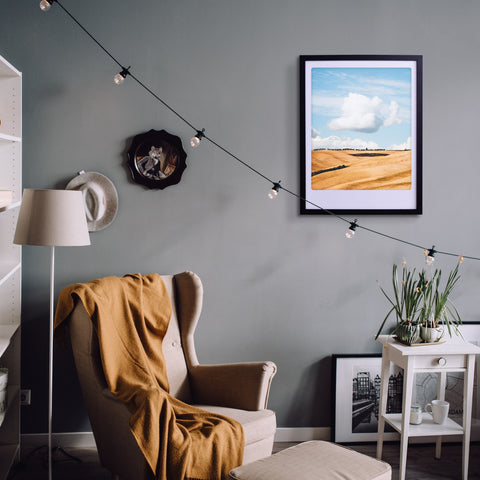 Gemütliches Wohnzimmer mit Sessel, Lampe, Pickmotion's "soft cloud" Art Poster in einem maßgefertigten Bilderrahmen, Lichterketten und Pflanzen vor einer grauen Wand für stilvollen Charakter.