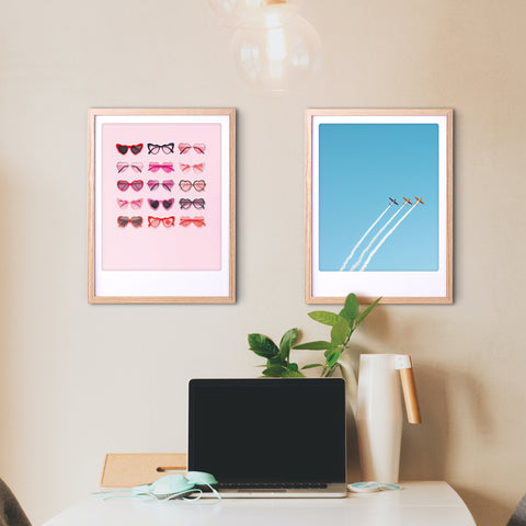 Ein minimalistischer Schreibtisch mit Laptop und Pflanze, darüber zwei Pickmotion-Plakate zum großen Geburtstag - Sonnenbrillen auf rosa und Flugzeuge im blauen Himmel - verleihen dem Raum einen stilvollen Charakter.