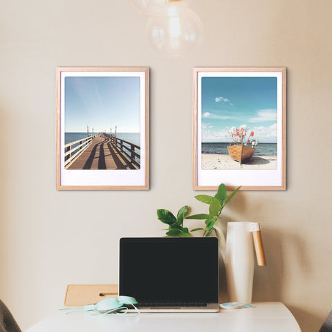 Ein Schreibtisch mit einem Laptop, einer Pflanze und zwei Pickmotion "Auszeit"-Kunstpostern in maßgefertigten Rahmen, die an der beigen Wand darüber hängen.