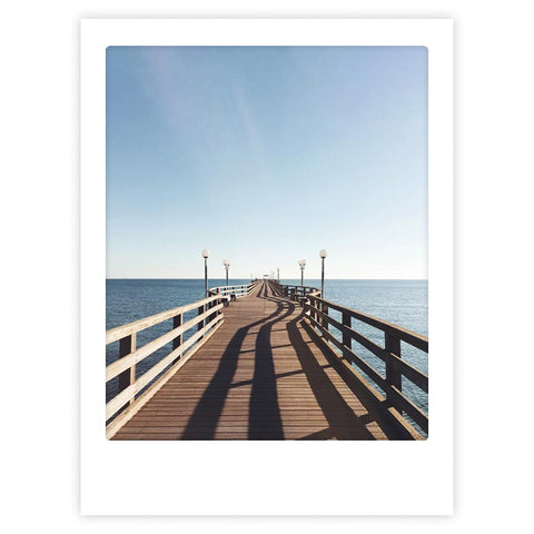 Das "seebrucke" Kunstposter von Pickmotion zeigt eine hölzerne Seebrücke, die sich über das ruhige blaue Wasser erstreckt, mit Laternen entlang des Weges, und ist ideal für die Präsentation in individuellen Rahmen oder als stilvolle Wanddekoration.