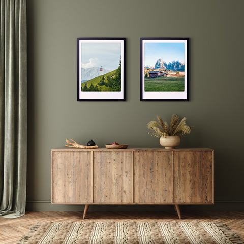 Ein hölzernes Sideboard mit Dekor sitzt unter zwei bergauf Bilderrahmen von Pickmotion, die Landschaftsfotos auf einer olivgrünen Wand präsentieren und dem Raum einen stilvollen Charakter verleihen.