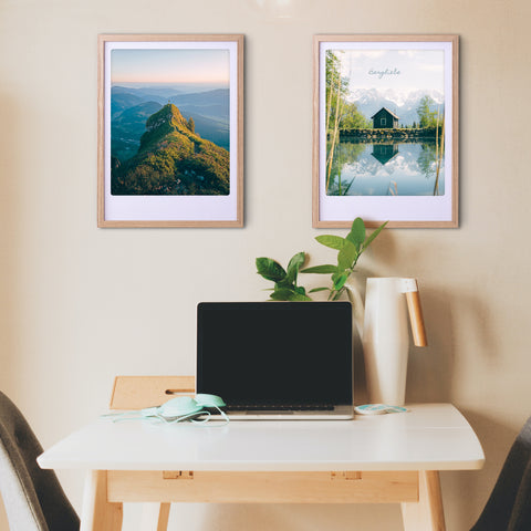 Ein minimalistisch eingerichteter Schreibtisch mit Laptop, Pflanze und zwei maßgefertigten Pickmotion Bergspiegel-Bilderrahmen mit Art-Poster-Drucken an der Wand darüber, die dem Raum stilvollen Charakter verleihen.