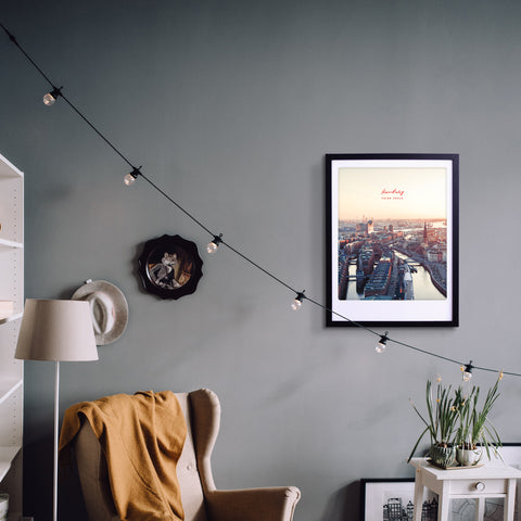 Gemütliche Wohnzimmerecke mit Sessel, Lampe, Pickmotion-Kunstposter "Sonnenuntergang über Hamburg" im Rahmen, Stadtfoto und Lichterkette an der Wand für stilvollen Charakter.