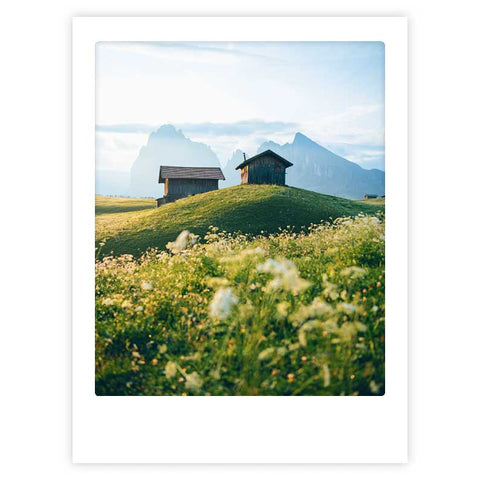 Zwei Holzhütten auf einem grasbewachsenen Hügel mit Wildblumen und nebligen Bergen im Hintergrund - "the sound of music" Art Poster von Pickmotion. Ideal für individuelle Rahmen und elegante Innenräume, die den stilvollen Charme der Natur einfangen.