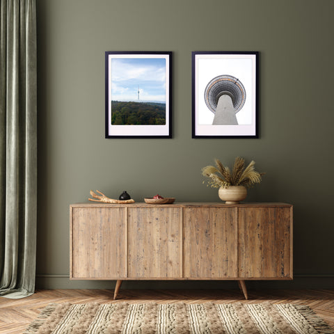 Holzanrichte mit Dekor unter zwei maßgefertigten Bilderrahmen, die jeweils die "blick nach oben"-Kunstposterdrucke von Pickmotion mit einem hohen Turm auf einer grünen Wand für stilvollen Charakter zeigen.