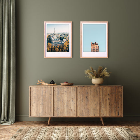 In einem gemütlichen Zimmer steht ein hölzernes Sideboard mit Dekoration unter zwei gerahmten "autumn in paris" Kunstpostern von Pickmotion an einer olivgrünen Wand.