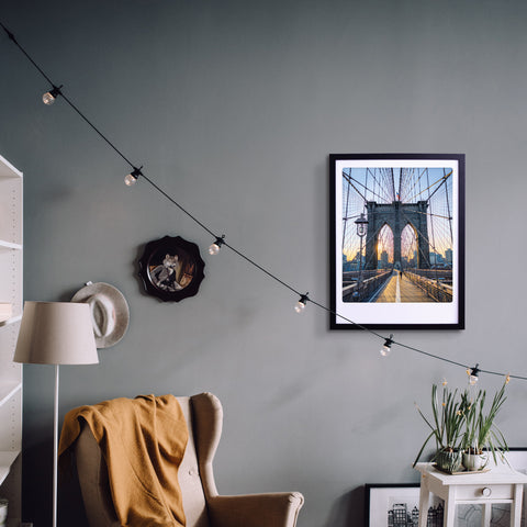 Gemütliche Wohnzimmerecke mit einem beigen Sessel, Pflanzen und dem Pickmotion "walk with me to nyc" Kunstposter in einem maßgefertigten Bilderrahmen an einer grauen Wand, die stilvollen Charakter in den Raum bringt.