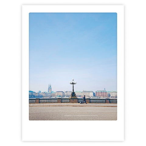 Eine Person geht an einem Laternenpfahl auf einer Brücke mit städtischen Gebäuden und blauem Himmel vorbei - Asterspaziergang von Pickmotion, ideal als Instagram-Motiv oder auffälliges Kunstposter für Ihren individuellen Bilderrahmen.