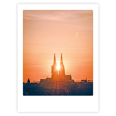 Sunset in cologne von Pickmotion-ein Kunstposter mit der Silhouette einer Kathedrale bei Sonnenuntergang, bei der das Sonnenlicht zwischen den Zwillingstürmen hindurchscheint. Stilvoller Charakter, ideal für maßgefertigte Bilderrahmen.