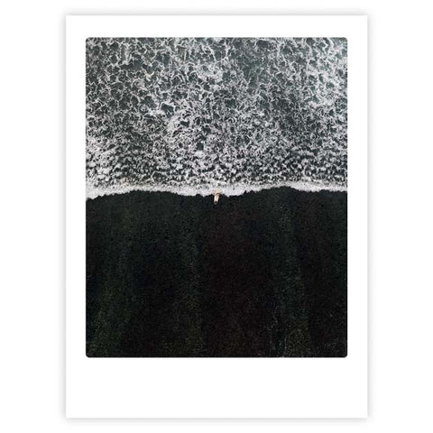 Luftaufnahme einer Person am Rande der schäumenden Wellen an einem dunklen Sandstrand - "Black Beach" von Pickmotion, perfekt als Kunstposter, das von internationalen Instagram-Fans geliebt wird.