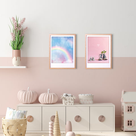 Ein pastellfarbenes Kinderzimmer mit dem Kunstposter "a rainbow in the sky" von Pickmotion in einem Rahmen, gepaart mit Regenbogen- und Schuh-Wandbildern, rosa-weißem Dekor und Blumen auf einem Regal.