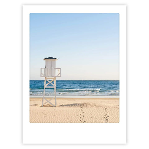 Das Kunstposter "a morning at the beach" von Pickmotion zeigt einen weißen Rettungsschwimmerturm am Sandstrand mit ruhigen Wellen und einem klaren Himmel - ideal für maßgefertigte Bilderrahmen.
