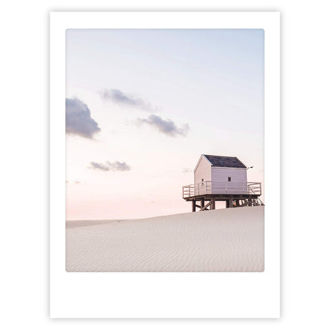 Das Kunstposter "the house by the ocean" von Pickmotion zeigt ein kleines weißes Haus auf Stelzen, das allein an einem Sandstrand unter einem pastellfarbenen Himmel steht und jedem Raum einen stilvollen Charakter verleiht.
