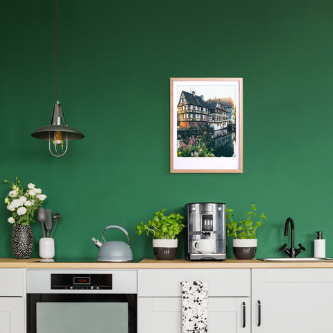 Eine moderne Küche mit grünen Wänden, Pflanzen, einer Kaffeemaschine und dem "Sommerabend in Straßburg" Kunstposter von Pickmotion in einem maßgefertigten Bilderrahmen für stilvollen Charakter über dem Tresen.