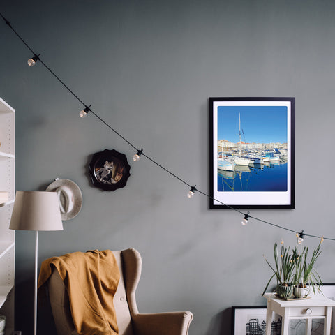 Gemütliches Zimmer mit Sessel, Lampe, Pflanzen und einem stilvollen Kunstposter in einem maßgefertigtem Bilderrahmen. Das "by the harbour" gerahmte Foto von Pickmotion und Lichterketten verleihen der grauen Wand stilvollen Charakter.