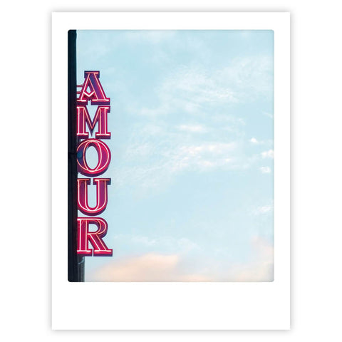 Das Pickmotion-Neonschild "amour" leuchtet auf diesem Kunstposter im Polaroid-Stil rosa vor einem wolkenverhangenen blauen Himmel und verleiht jedem Raum einen stilvollen Charakter.