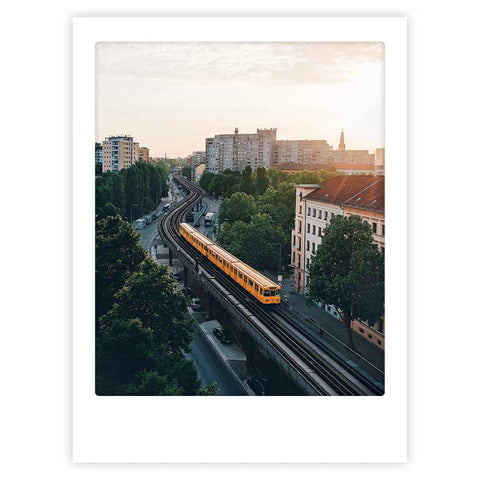 Durch die Stadt" von Pickmotion zeigt einen gelben Zug mit stilvollem Charakter, der bei Sonnenuntergang zwischen Bäumen und Gebäuden über erhöhte Gleise gleitet - ein ideales Kunstposter, um Ihren Raum zu verschönern.