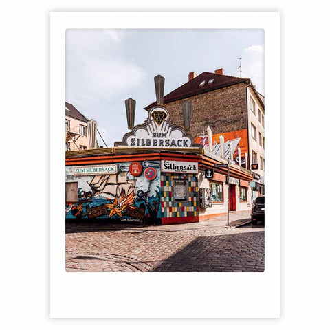 Das "City Corner"-Kunstposter von Pickmotion zeigt Zum Silbersack, ein lebhaftes Café mit Wandmalerei und stilvollem Charakter an einer Straßenecke mit Kopfsteinpflaster, das jeden Raum aufwertet.