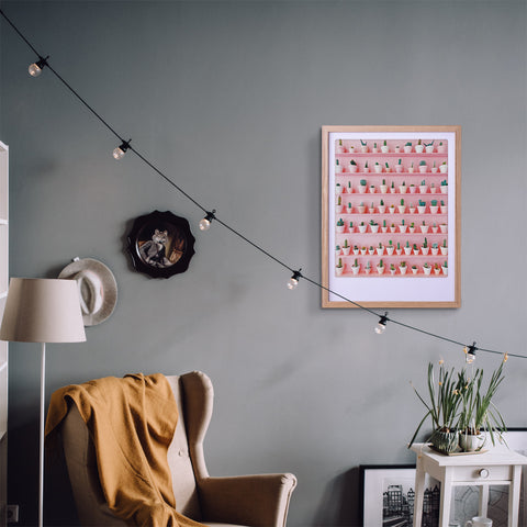 Gemütliches Wohnzimmer mit einem Sessel, gelber Decke, Lichterketten und einem Pickmotion-Kaktus-Wandkunstposter in einem Bilderrahmen an der grauen Wand.