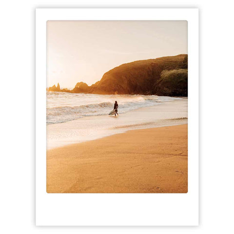Ein einsamer Surfer spaziert bei Sonnenuntergang an einem Sandstrand entlang, mit Wellen und Klippen im Hintergrund - ideal als "Surfer am Strand" Kunstposter von Pickmotion, um internationales Design-Flair in Ihren Raum zu bringen.