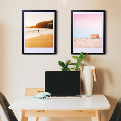 Ein Schreibtisch mit Laptop, Pflanze und zwei Pickmotion "Strandhaus" Art Poster Drucke in maßgefertige Bilderrahmen an der Wand darüber.