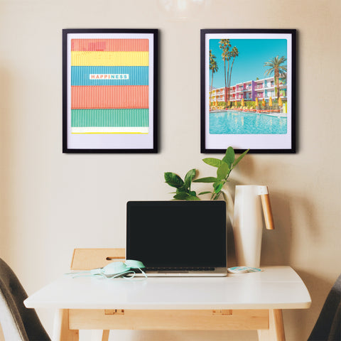Bunte Behälter von Pickmotion verleihen einem weißen Schreibtisch mit Kunstdrucken, Pflanze und Krug vor einer beigen Wand stilvollen Charakter und kuratiertes Art-Poster-Flair.