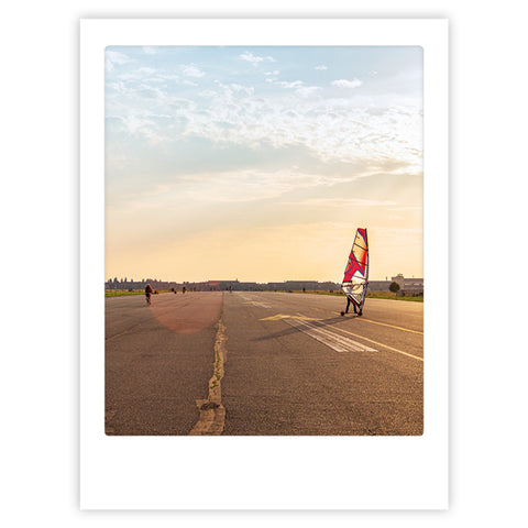 Eine Person beim Windsurfen auf dem leeren Tempelhofer Feld bei Sonnenuntergang mit einem bunten Segel und wolkenverhangenem Himmel - eine ikonische Szene voller stilvoller Charakter, perfekt als Art Poster von Pickmotion.