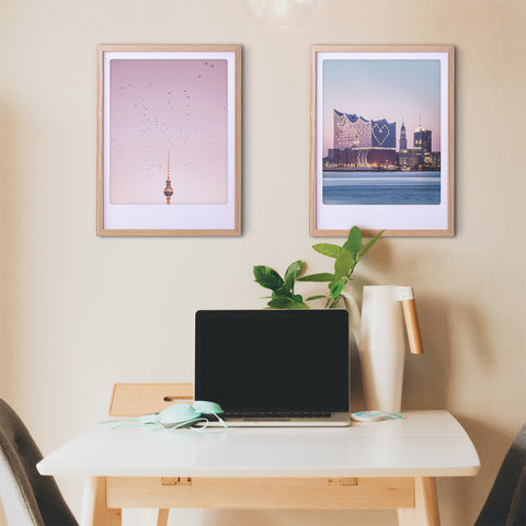 Minimalistischer Arbeitsbereich mit einem Laptop auf einem weißen Schreibtisch, zwei Pickmotion maßgefertigte Bilderrahmen mit dem "birds above berlin" Art Poster, und eine Pflanze an einer beigen Wand für stilvollen Charakter.