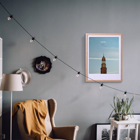 Ein gemütliches Wohnzimmer mit Sessel, gelber Decke, Lichterkette, Topfpflanze und einem Pickmotion-Kunstposter der St. Michaelis Kirche in einem exklusiv maßgefertigten Bilderrahmen.