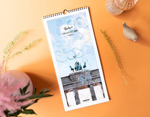 Ein Berliner Wandkalender mit Sprechblasen, auf einer orangefarbenen Fläche mit Vasen, einer Pflanze und Trockenblumen.