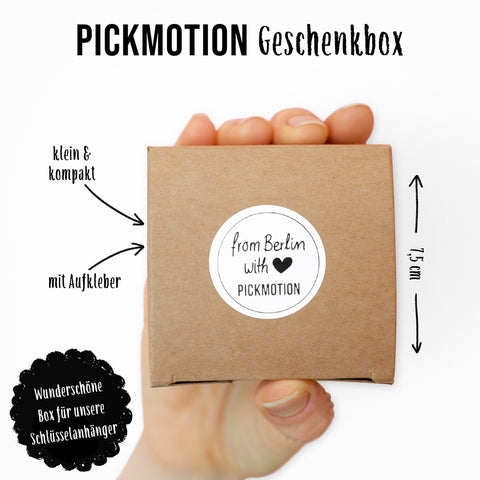 Eine Hand hält eine kleine braune Geschenkschachtel mit der Aufschrift "beste Schwester" von Pickmotion, mit deutschem Text und Maßangaben. Im Inneren befindet sich ein Edelstahl Schlüsselanhänger mit elegantem Reliefdruck.