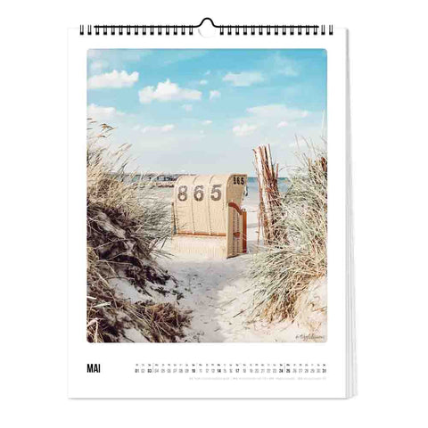 Ein Strandkorb mit der Aufschrift 865 sitzt im Sand zwischen Gräsern unter einem blauen Himmel - ideal für Liebhaber des Küste & Meer - Kalender (Format 30x40) von Pickmotion.