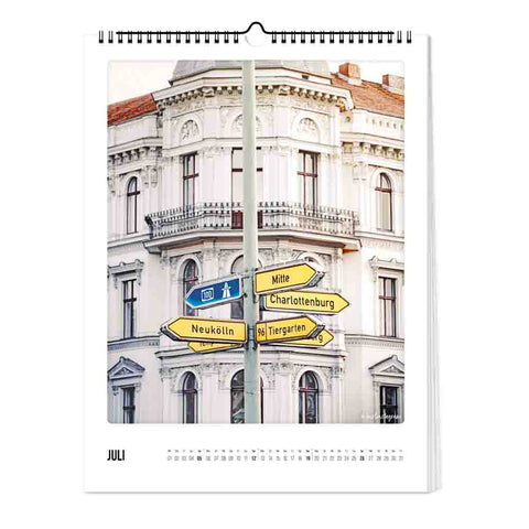 Straßenschilder zu Berliner Bezirken stehen vor einem weißen Gebäude auf der "Juli"-Seite des Berlin-Kalenders (Format 30x40) von Pickmotion - ideal für Fans von minimalistischem Design in ihrer Wandkalender-Kollektion.