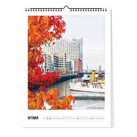 Eine Uferszene mit herbstlichen Bäumen, einem angedockten Boot und modernen Gebäuden - abgebildet im Hamburg - Kalender (Format 30x40) von Pickmotion, ideal für jeden Fotokalender mit nordischem Design.