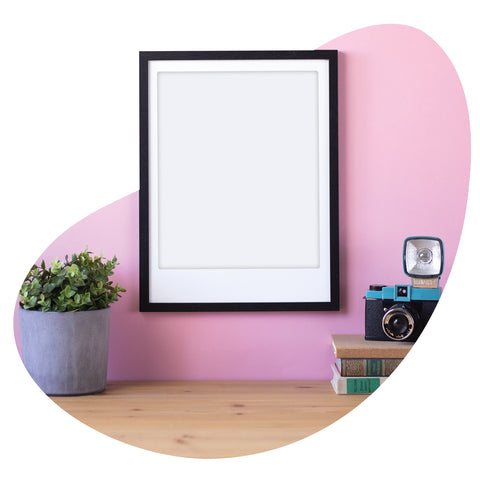 Artposter von Pickmotion, ein alter Fotoapparat, gestapelte Bücher und eine Topfpflanze stehen auf einem Holztisch vor einer rosafarbenen Wand, die Produkte aus früheren Releases in stilvoller Harmonie präsentiert.