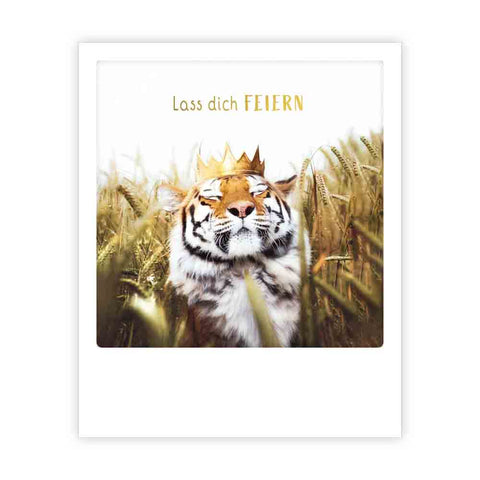 Ein gekrönter Tiger sitzt im hohen Gras, eingefangen im Sofortbild-Stil, mit dem Spruch "lass dich feiern" darüber-ideal für trendige Postkarten von Pickmotion oder als Inspiration für Instagram-Fotograf*innen.
