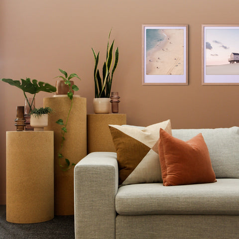Modernes Wohnzimmer mit einem neutralen Sofa, orangefarbenen Kissen, Topfpflanzen und dem "Strandtag"-Kunstposter von Pickmotion in maßgefertige Bilderrahmen für stilvollen Charakter.