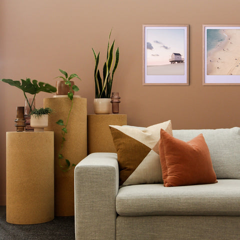 Modernes Wohnzimmer mit Topfpflanzen, beigem Sofa, rostfarbenen Kissen und dem Kunstposter "Das Haus am Meer" von Pickmotion in einem maßgefertigten Rahmen an einer hellbraunen Wand für stilvollen Charakter.