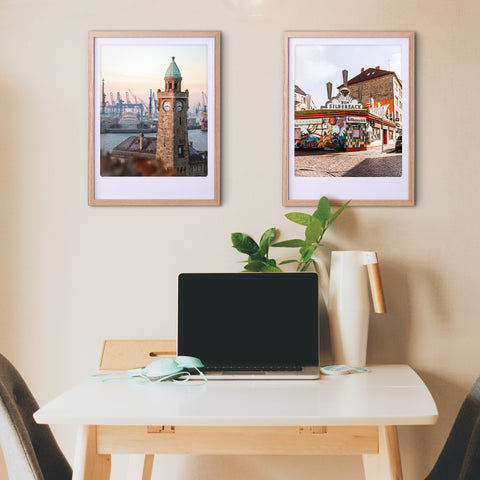 Minimalistischer Arbeitsbereich mit Laptop, Kopfhörern, Pflanze und zwei Pickmotion "city corner" Kunstpostern in maßgefertigten Bilderrahmen über dem Schreibtisch, die dem Raum einen stilvollen Charakter verleihen.