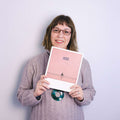 Frau mit Brille lächelt und hält ein rosa Buch mit einer kleinen Illustration auf dem Einband.