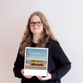Eine Frau mit Brille hält ein gerahmtes Foto eines gelben Lieferwagens am Meer, auf dem "Oh happy day" steht.
