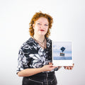 Eine Person mit lockigem Haar hält ein Foto eines auf dem Wasser schwimmenden Hauses mit schwarzen Luftballons vor einem blauen Himmel.