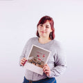 Frau mit roten Haaren, die ein Foto mit Büchern und Blumen in der Hand hält, vor einem schlichten weißen Hintergrund.