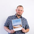 Ein Mann in einem geblümten Hemd hält ein Foto einer Strandszene mit Text vor einem schlichten weißen Hintergrund.