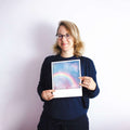 Frau mit Brille, die ein Foto eines Regenbogens gegen den Himmel hält, stehend vor einem weißen Hintergrund.