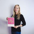 Frau mit langen blonden Haaren hält ein rosa Schild mit der Aufschrift BE HAPPY vor einem schlichten hellen Hintergrund.